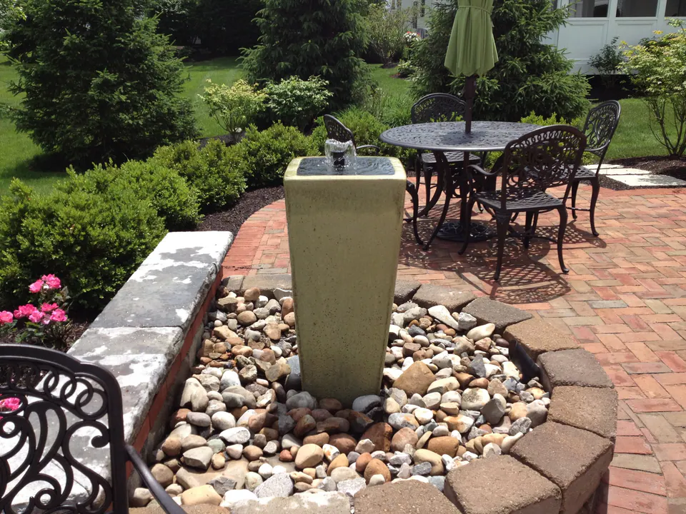 Tall urn fountain in a river-rock bed at the edge of a brick patio with dining table